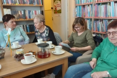 Kobiety siedzą wokół stołu, na którym stoją filiżanki z herbatą,  i przeglądają zdjęcia