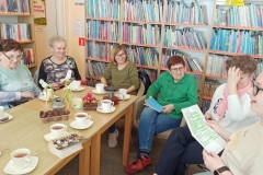 Kobiety siedzą wokół stołu, na którym stoją filiżanki z herbatą,  i przeglądają zdjęcia i pamiątki