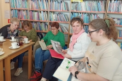 Kobiety siedzą wokół stołu, na którym stoją filiżanki z herbatą,  i przeglądają zdjęcia i pamiątki